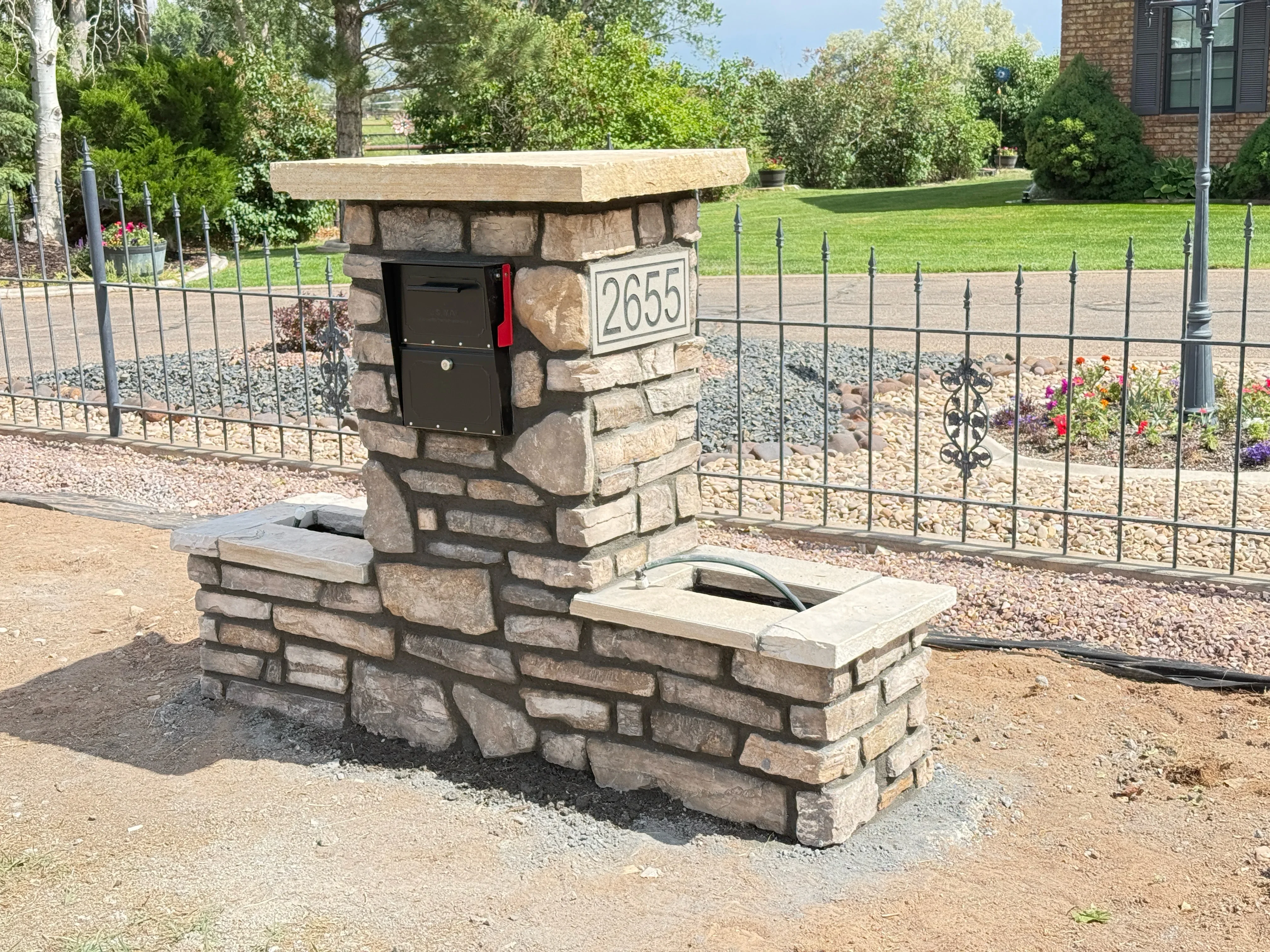 Stacked stone mailbox post with black mailbox and address plaque, concrete cap, gravel base