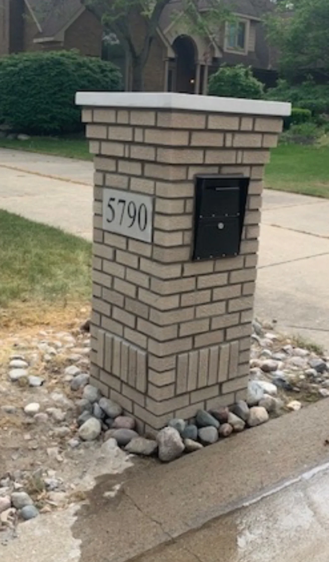 Tan brick mailbox with white cap stone, black mailbox door, and river rock base