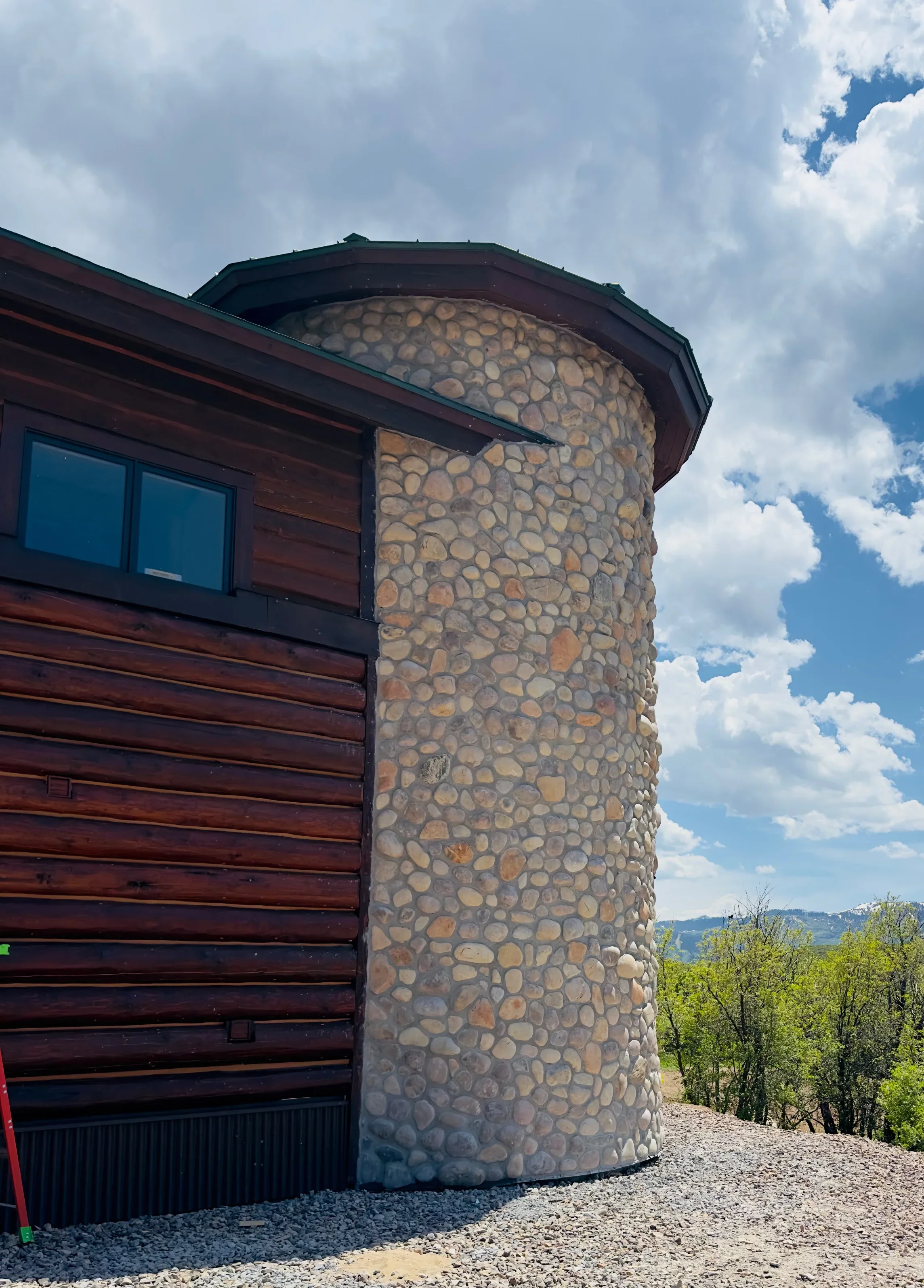 Cylindrical stone tower with stacked river rock masonry and metal roof cap beside log cabin